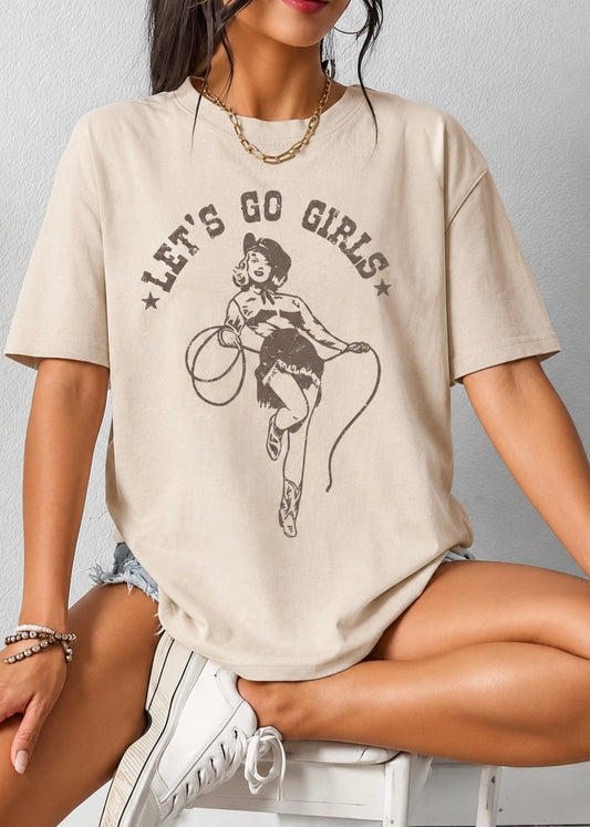 A woman wearing an oversized beige graphic t-shirt with a retro pin-up illustration and the text 'LET'S GO GIRLS' printed on the front, paired with denim shorts and white sneakers.