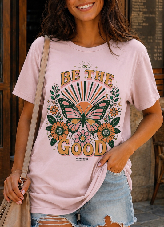 Person wearing a pink t-shirt with a butterfly graphic and 'Be the Good' text.