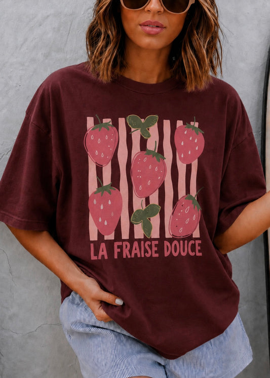 A woman wearing an oversized maroon graphic t-shirt with a strawberry print and the French text 'LA FRAISE DOUCE' on the front, paired with light blue denim shorts against a gray concrete wall.