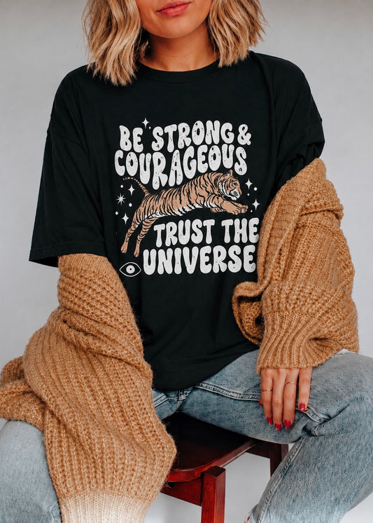 A woman wearing a black graphic t-shirt with a white text and tiger design, styled with a chunky brown knit cardigan and light wash jeans, sitting on a wooden stool.