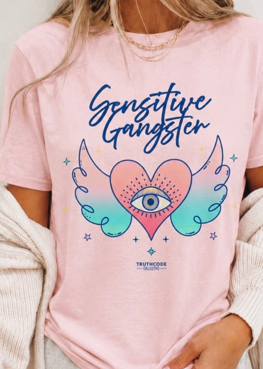 Person wearing a pink t-shirt with 'Sensitive Gangster' design and 'Truthcode Collective' branding. Graphic shows all seeing eye, and heart with wings.