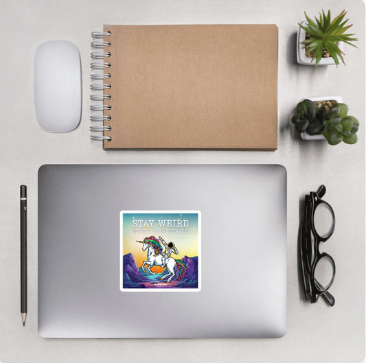 Aerial view of a desktop with laptop showing a squared sticker of an astronaut riding a unicorn and text 'Stay weird'