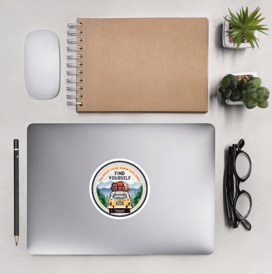 Aerial view of a desktop with laptop showing a circular sticker with design of a car packed with luggage and text 'Unleash your inner explorer' and 'FIND YOURSELF'