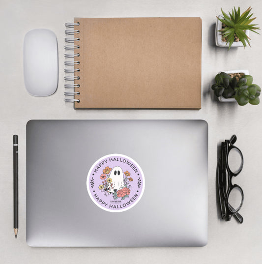 Aerial view of a desktop with laptop showing a circular sticker with cartoon ghost and floral design and text 'Happy Halloween'