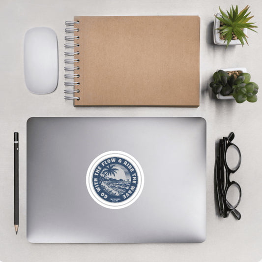 Aerial view of a desktop with laptop showing a circular sticker with a palm tree and ocean design and text 'Go with the flow & ride the wave'