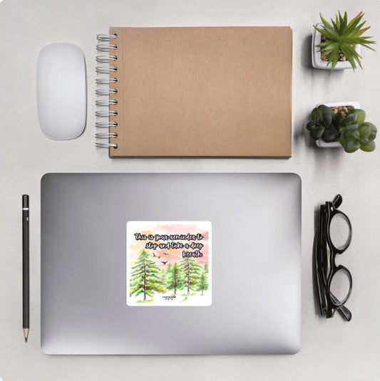 Aerial view of a desktop with laptop showing a squared sticker with a nature and pine tree design and text 'This is your reminder to stop and take a deep breath'