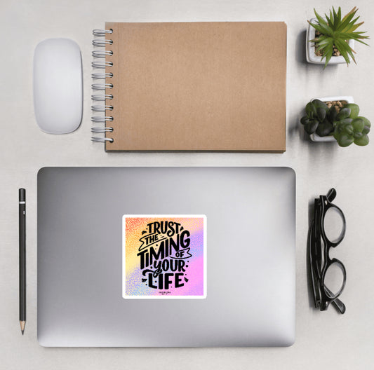 Aerial view of a desktop with laptop showing a colorful squared sticker with text 'Trust the timing of your life'