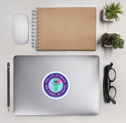 Aerial view of a desktop with laptop showing a circular sticker with a cute cartoon alien and text 'Hello human, we come in peace'