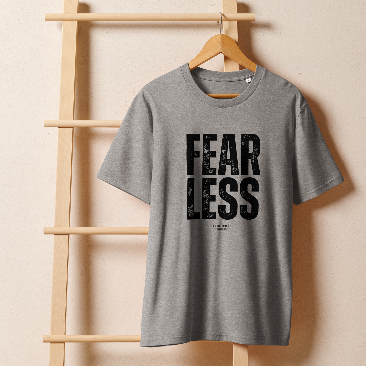 Gray organic t-shirt with 'FEAR LESS' text on a wooden hanger against a beige wall.