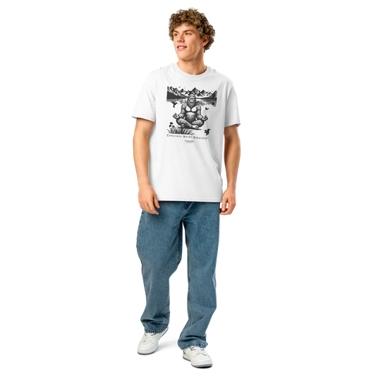 Guy wearing white organic t-shirt with a graphic of a meditating Sasquatch in a mountain setting and text reading 'Conscious, hairy, and unbothered'