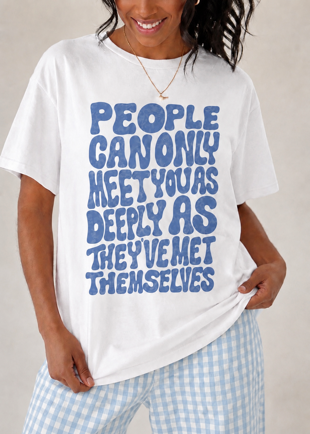 A woman wearing a white graphic t-shirt with blue text that reads 'PEOPLE CAN ONLY MEET YOU AS DEEPLY AS THEY'VE MET THEMSELVES', paired with light blue and white gingham pants.