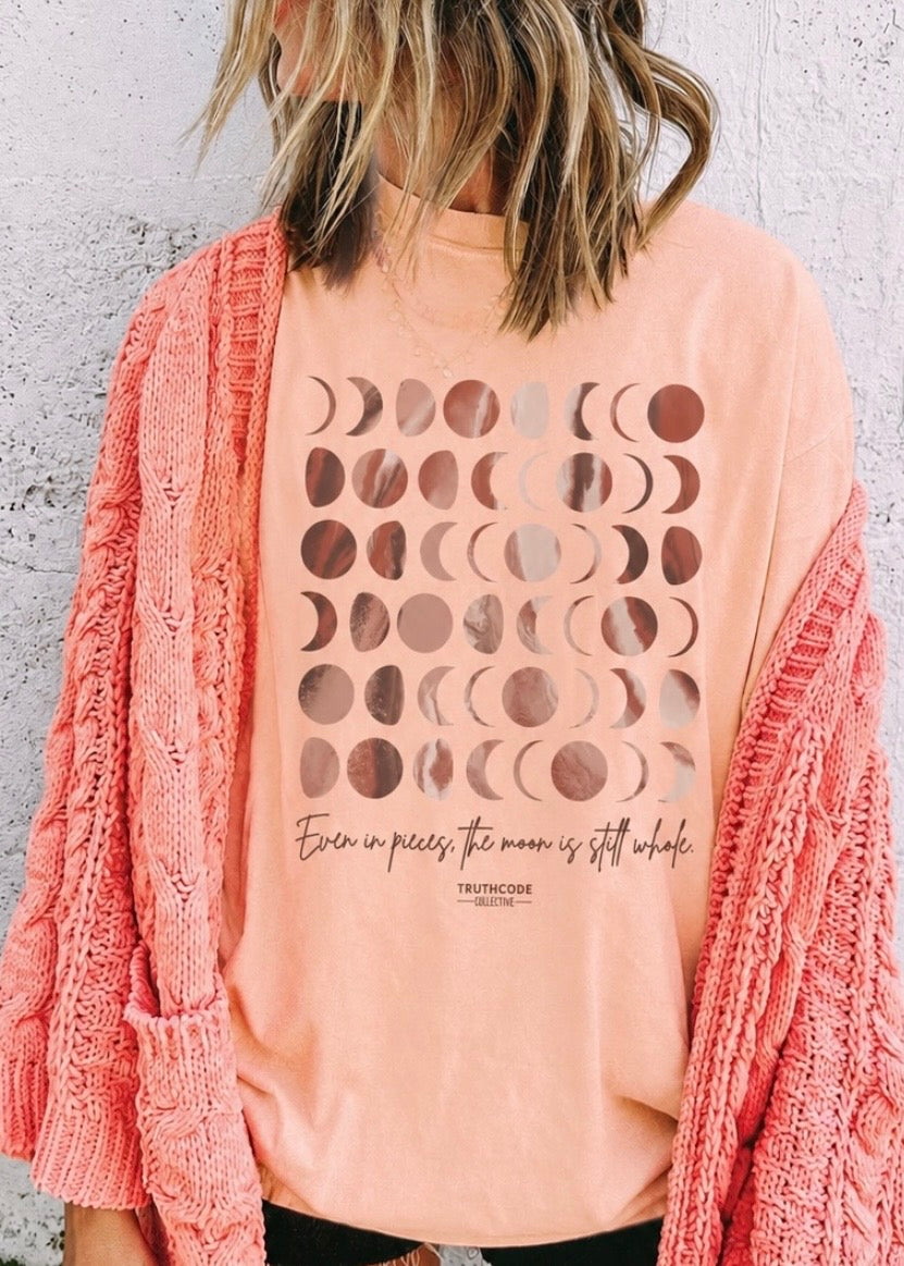 Person wearing a peach t-shirt with moon phase design and text, paired with a pink cardigan against a light gray background. Text says 'even in pieces the moon in still whole'.