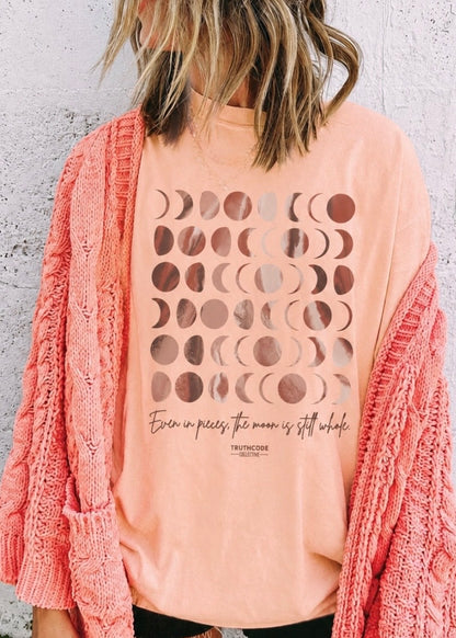Person wearing a peach t-shirt with moon phase design and text, paired with a pink cardigan against a light gray background. Text says 'even in pieces the moon in still whole'.