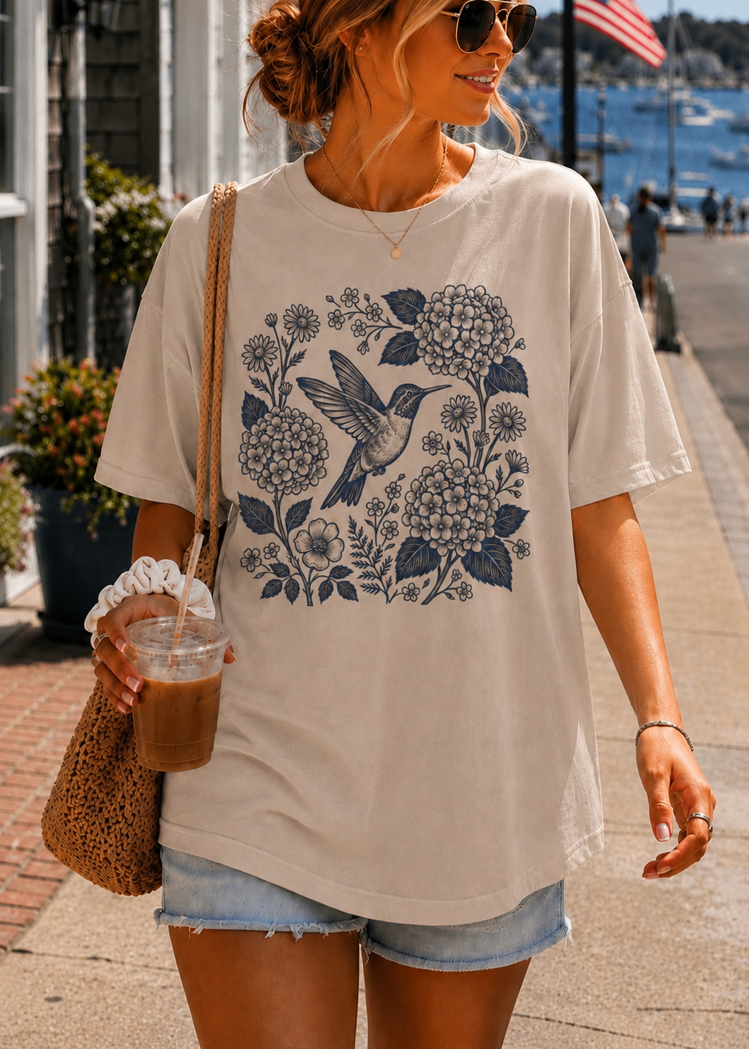 A woman wearing an oversized cream-colored t-shirt with a navy blue floral and hummingbird graphic, paired with denim shorts and sunglasses, holding an iced coffee on a sunny outdoor walkway.
