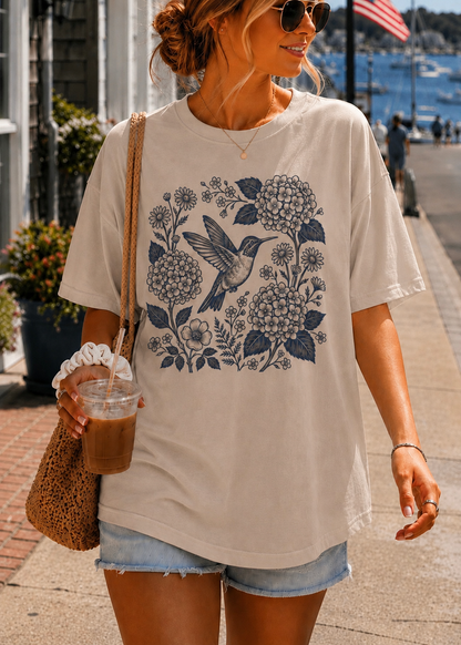 A woman wearing an oversized cream-colored t-shirt with a navy blue floral and hummingbird graphic, paired with denim shorts and sunglasses, holding an iced coffee on a sunny outdoor walkway.