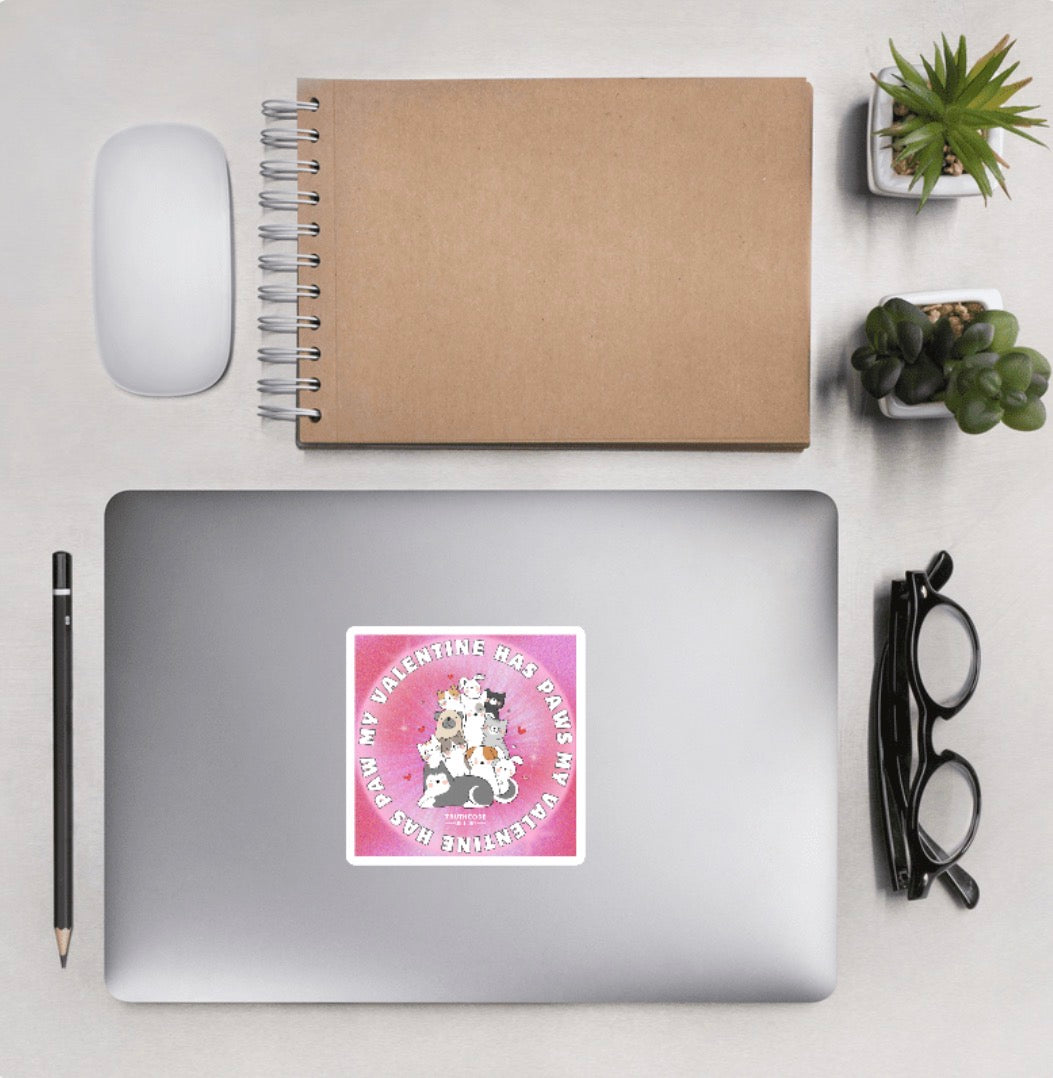Laptop featuring a pink circular sticker and text 'My valentine has paws' and graphics of dogs and cats, with desk accessories around.
