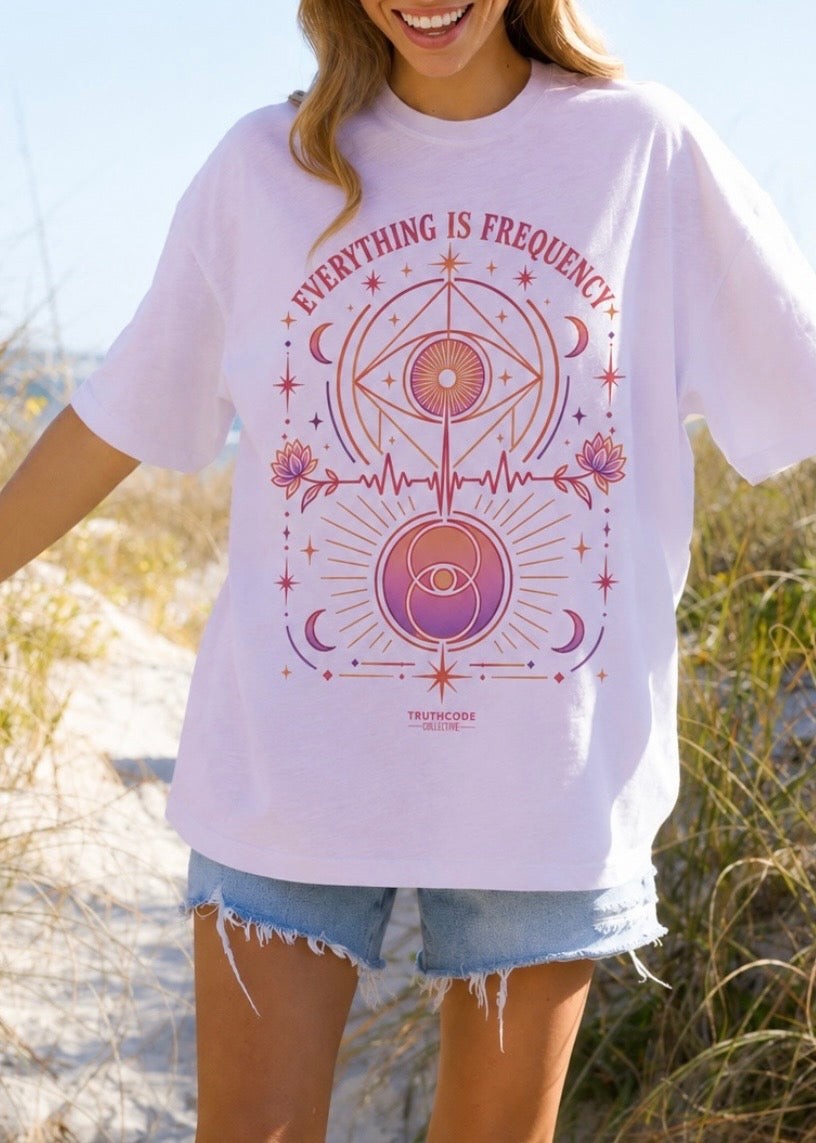 Person wearing a white t-shirt with a pink graphic design outdoors. Text says 'Everything is Frequency' and graphic includes all seeing eye, moons, stars, sacred geography, and lotus flowers.
