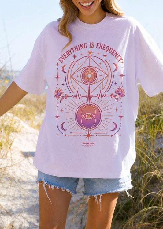Person wearing a white t-shirt with a pink graphic design outdoors. Text says 'Everything is Frequency' and graphic includes all seeing eye, moons, stars, sacred geography, and lotus flowers.