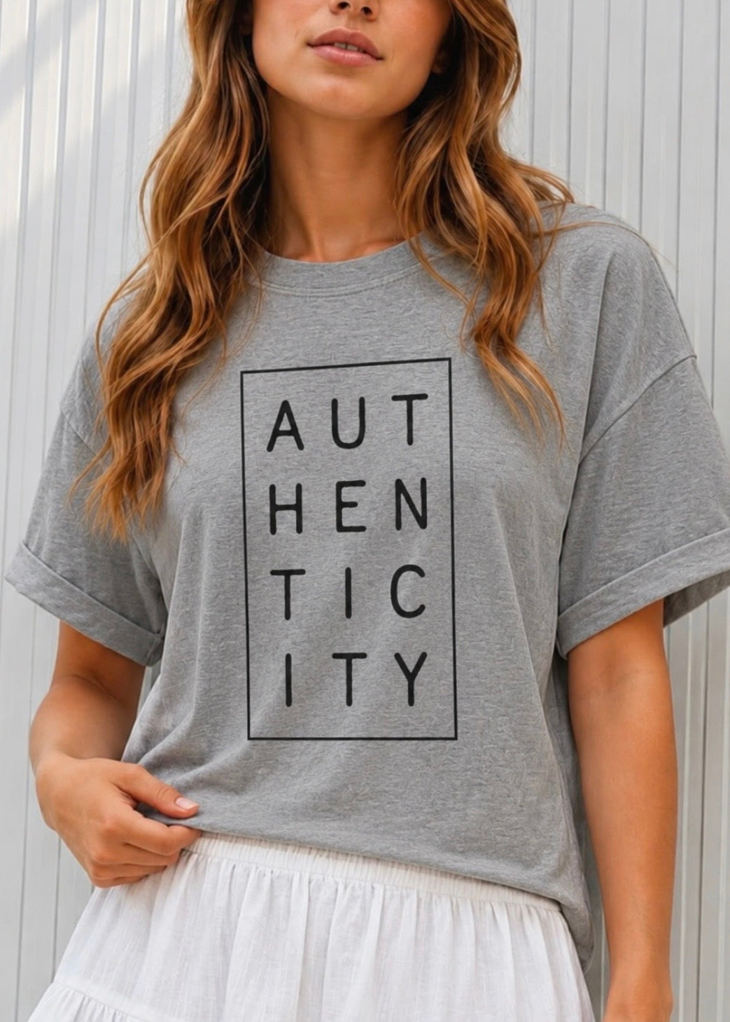 A woman wearing a heather gray oversized t-shirt with a black graphic print that reads 'AUTHENTICITY' inside a rectangular border, paired with a white skirt, set against a light gray paneled wall.