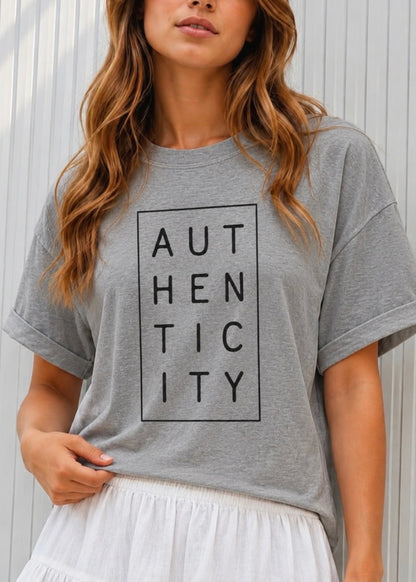 A woman wearing a heather gray oversized t-shirt with a black graphic print that reads 'AUTHENTICITY' inside a rectangular border, paired with a white skirt, set against a light gray paneled wall.