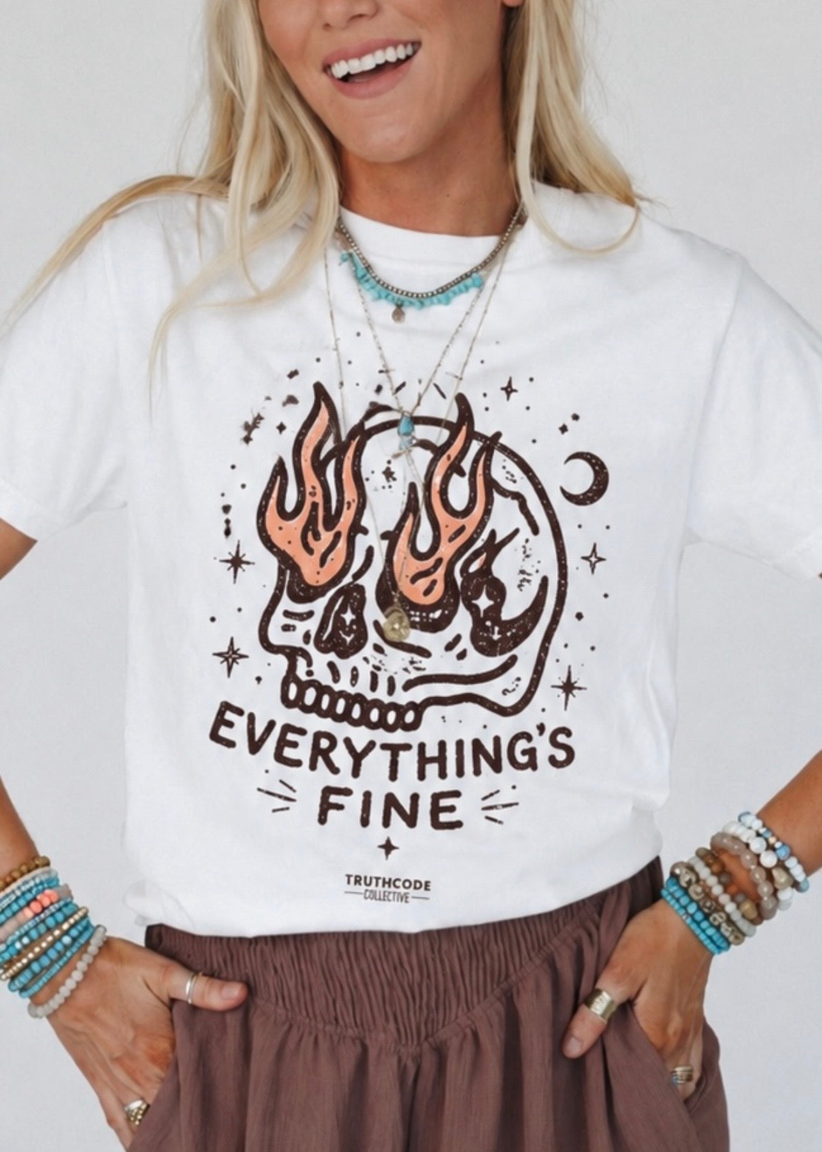 Person wearing a humorous white t-shirt with a skull with flames in its eyes graphic design and text 'EVERYTHING'S FINE' on a plain background. 