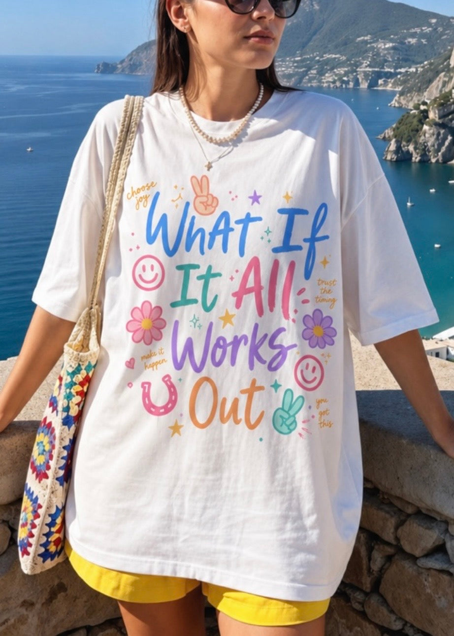 Person wearing a white t-shirt with colorful text and graphics, standing by a scenic coastal area. Text says 'what if t all works out'. Design includes peace signs, horse shoe, smiley faces, flowers and stars.