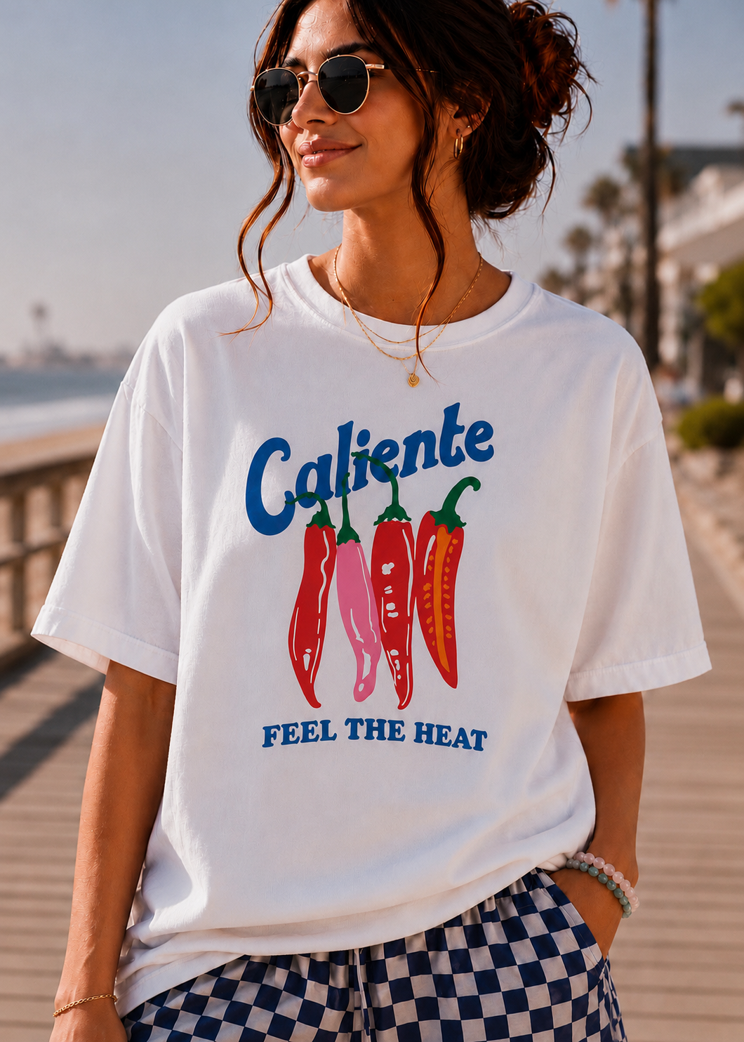 A woman wearing a white graphic t-shirt with 'Caliente' and 'FEEL THE HEAT' text above and below a graphic of four red and pink chili peppers, set against a sunny boardwalk background.