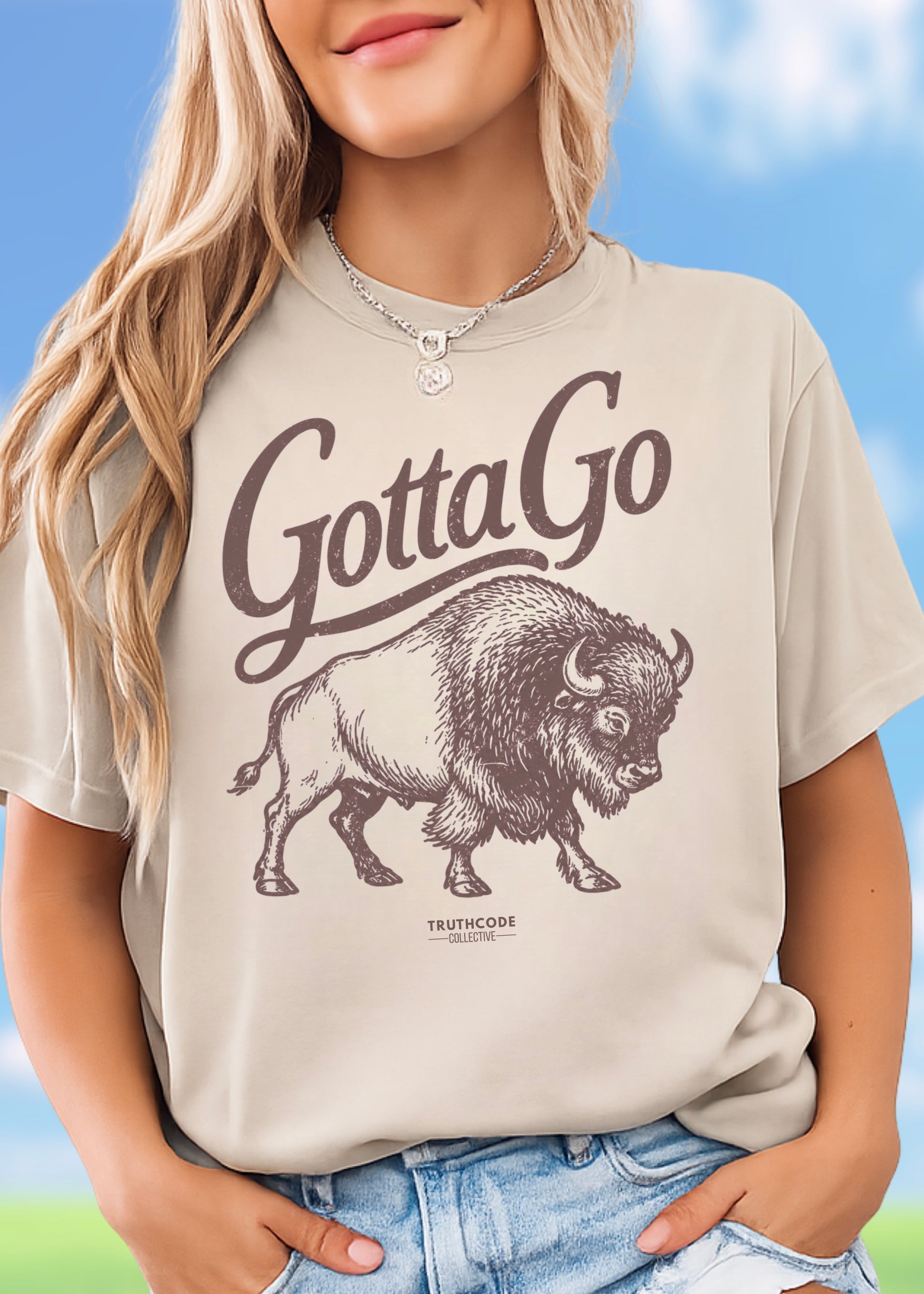 Person wearing a beige t-shirt with a graphic of a bison and 'Gotta Go' text, standing against a blue sky background.