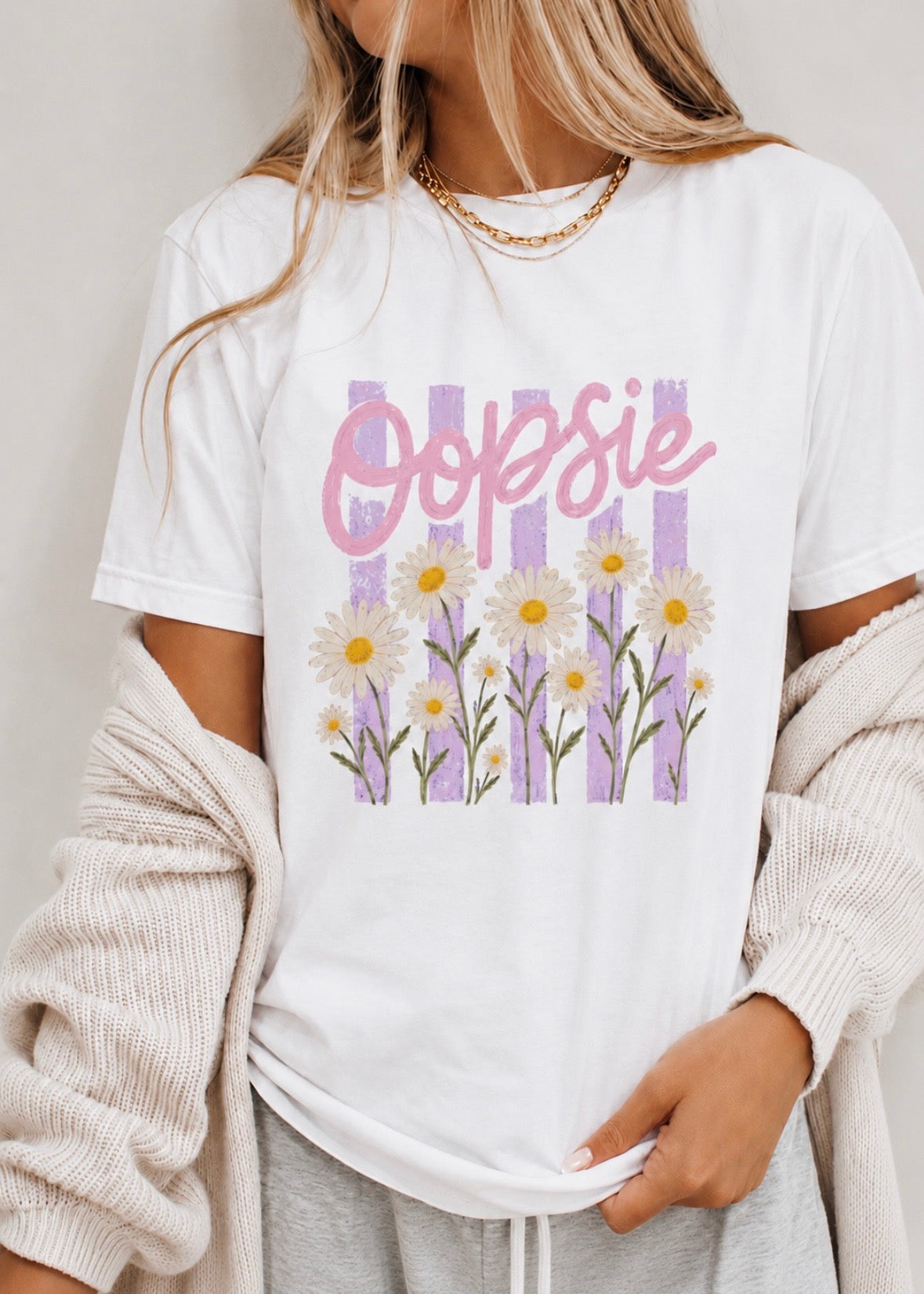 A woman wearing a white graphic t-shirt with a pink 'Oopsie' script and a purple floral daisy design, styled with a cream knit cardigan and gray sweatpants.