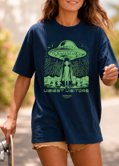 Person wearing a navy blue t-shirt with a green alien abduction graphic and text 'vibiest visitors'.