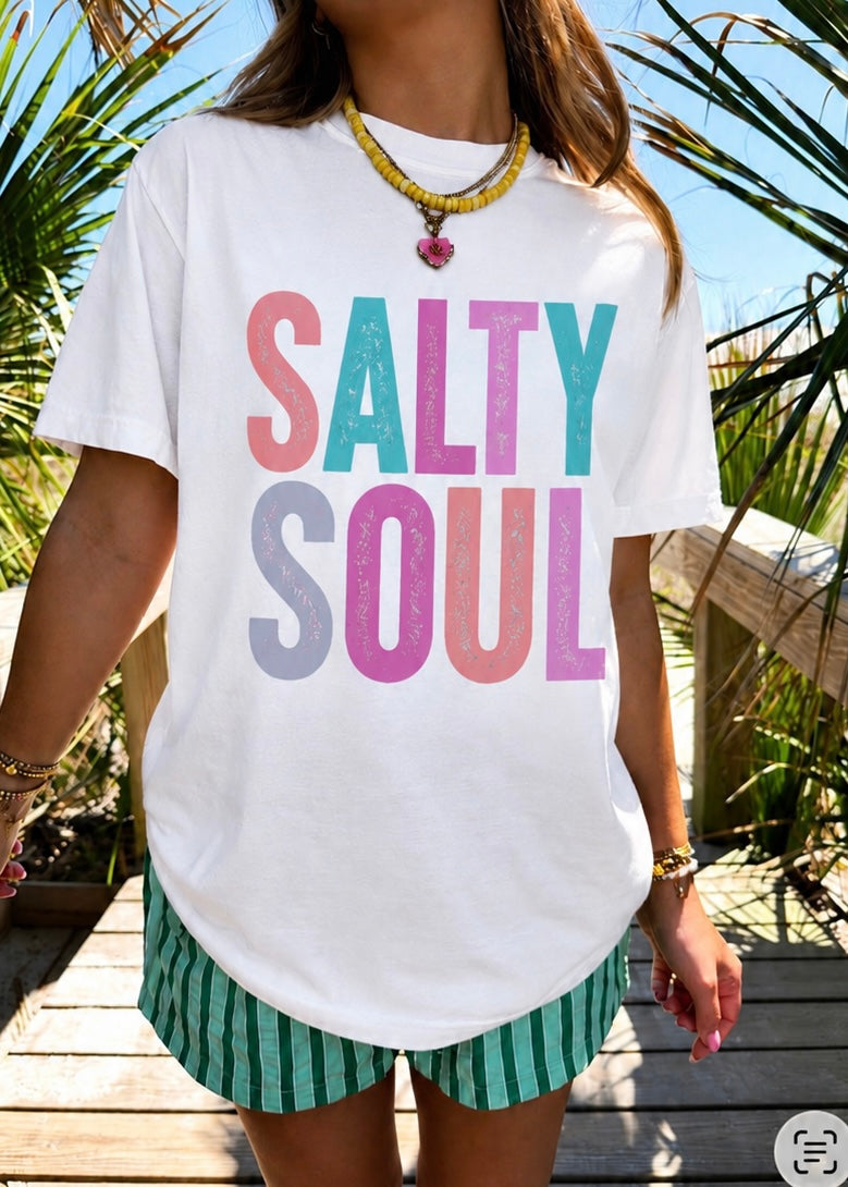 A woman wearing a white graphic t-shirt with 'SALTY SOUL' printed in pastel pink, teal, and lavender glitter-style letters, paired with green and white striped shorts, set against a sunny outdoor background with palm leaves.