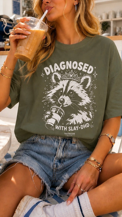 Person wearing a green t-shirt with a graphic design and text, holding a drink. Raccoon with tiara. Diagnosed with Slay-DHD.