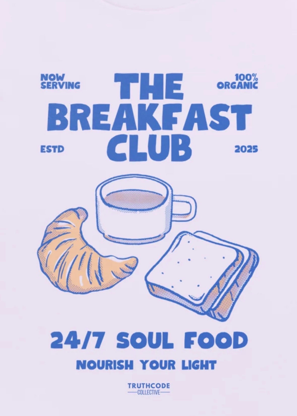 Close view of lavender t-shirt with text  'The Breakfast Club, 24/7 soul food, nourish the light ' with a cup of coffee, croissant, and toast