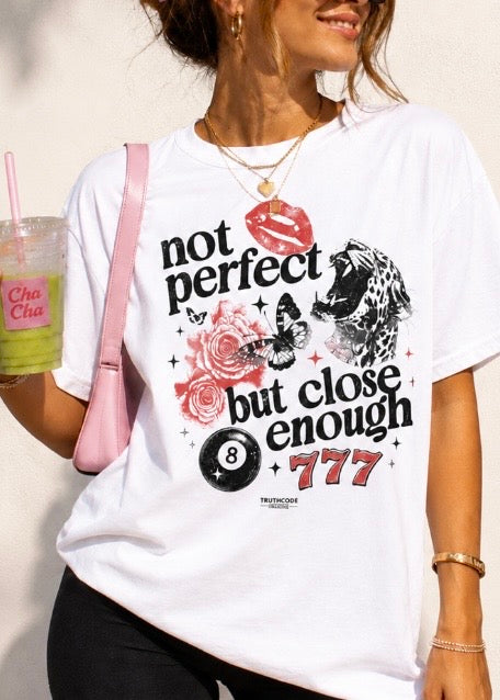 Person wearing a white t-shirt with graphic designs and text, holding a drink. Saying is not perfect but close enough. Includes tiger, butterfly, roses, eight ball, lips and 777.