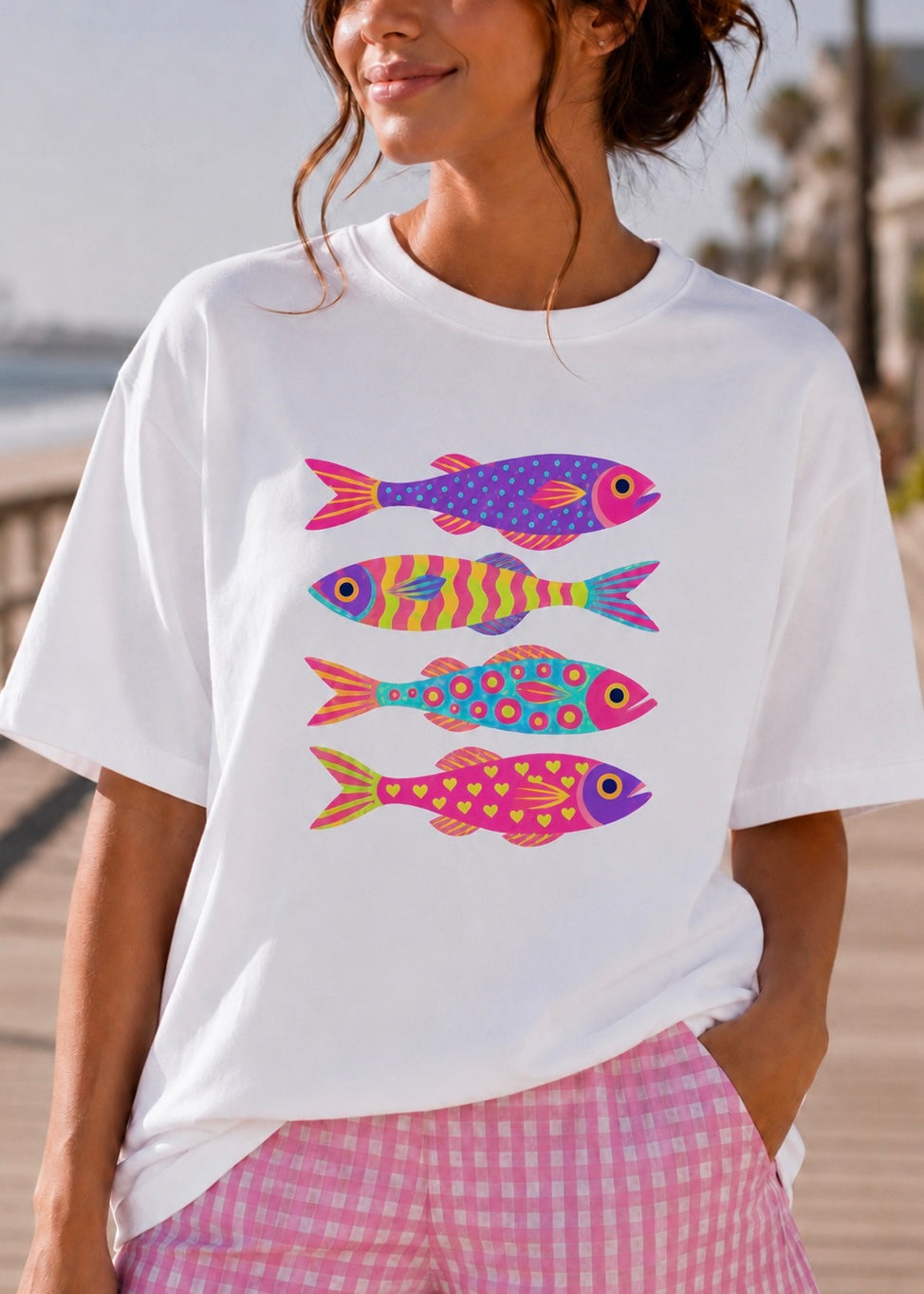 A woman wearing a white short-sleeve t-shirt with a colorful graphic of four stylized fish in pink, purple, orange, and blue, paired with pink and white gingham shorts.
