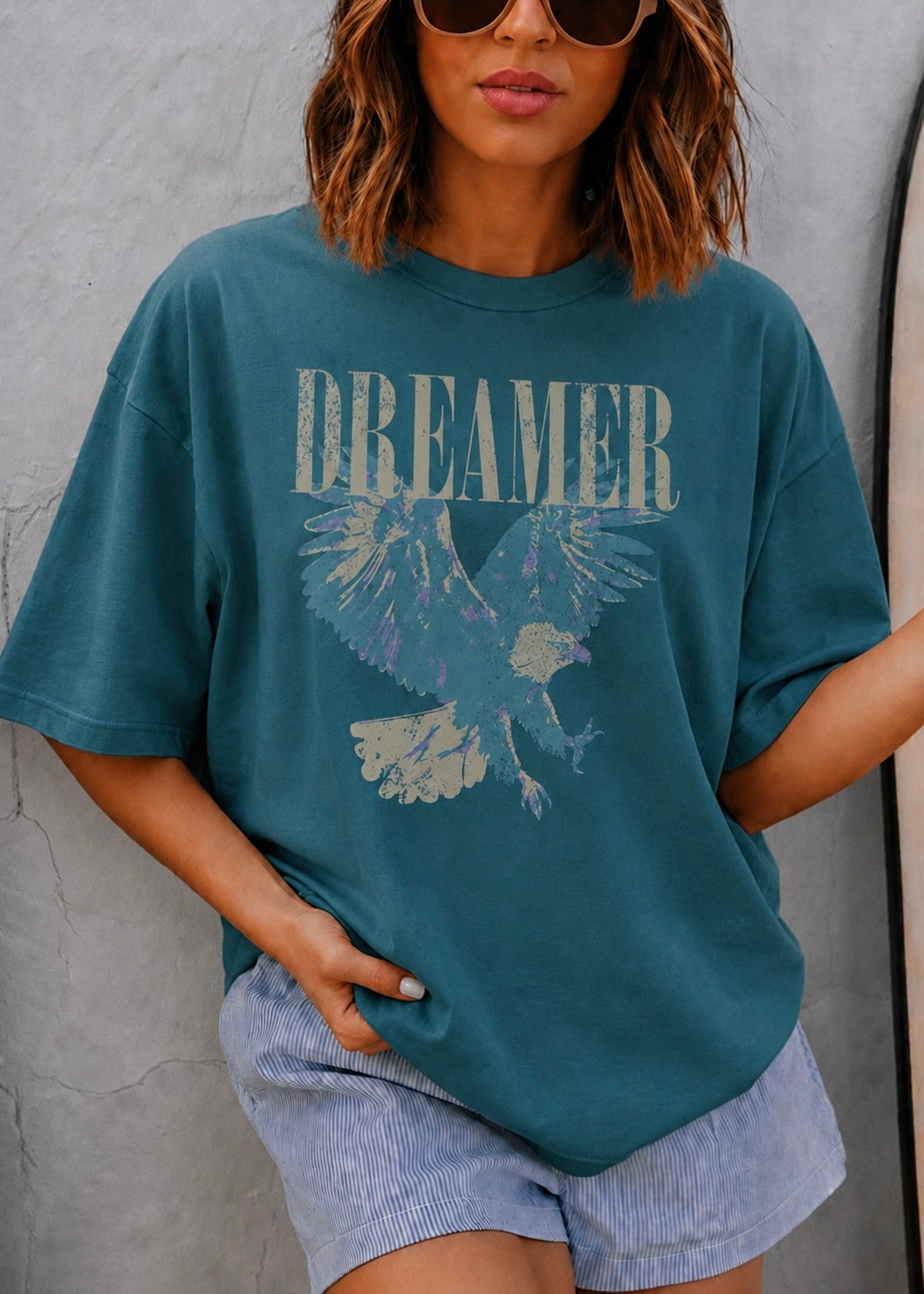 A woman wearing a teal blue oversized graphic t-shirt with a distressed 'DREAMER' text and eagle design, paired with light blue striped shorts, posing against a gray textured wall.
