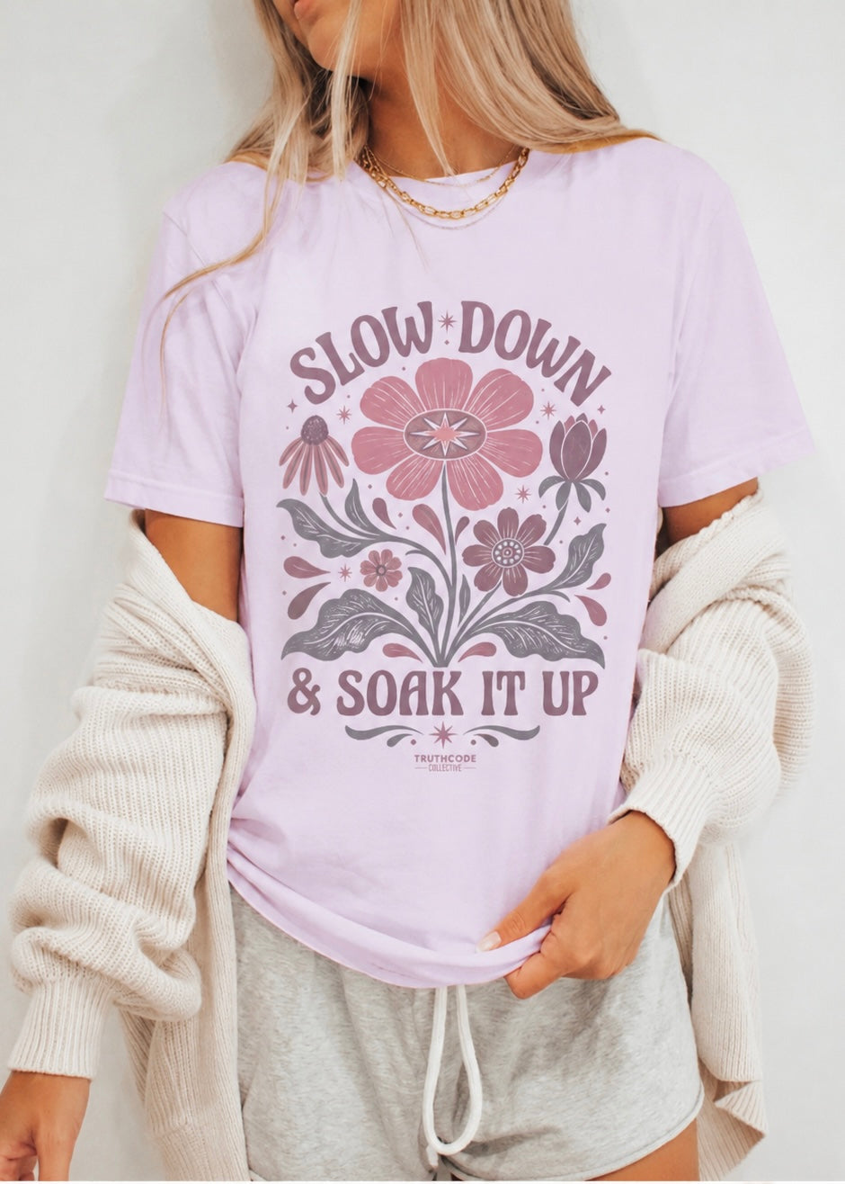 Person wearing a light purple t-shirt with floral design and text 'slow down and soak it up'.