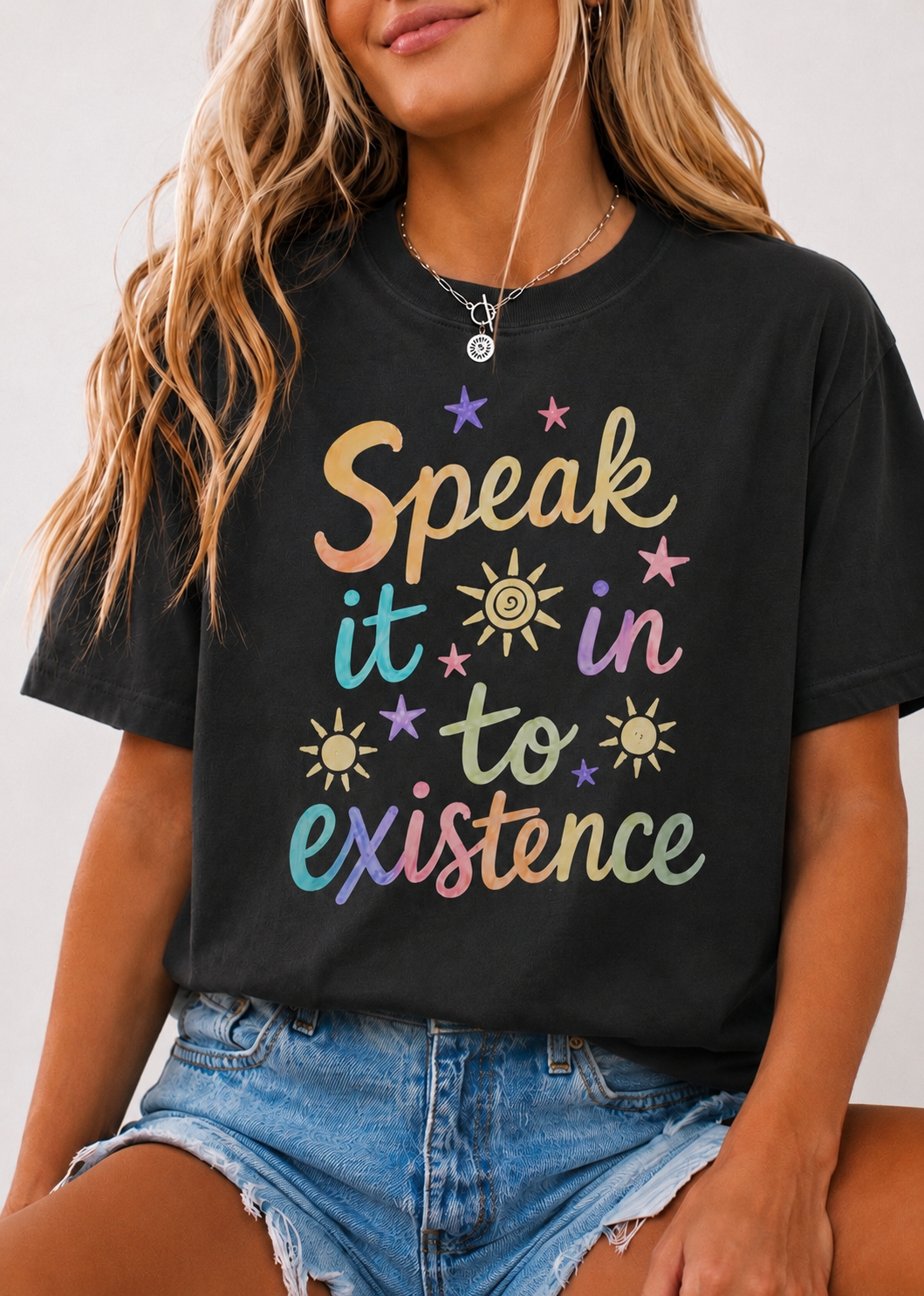 A woman wearing a black graphic t-shirt with the phrase 'Speak it in to existence' written in a colorful pastel gradient font, paired with denim shorts.