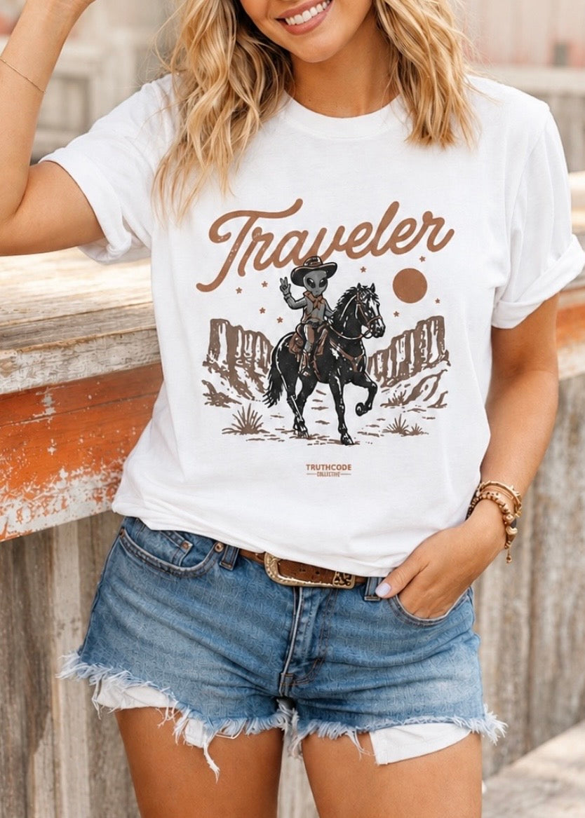 Person wearing a white t-shirt with a graphic design of alien holding up  peace sign and riding horse in desert and text that says 'traveler'. 