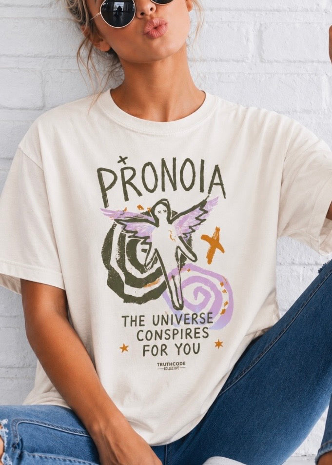 Person wearing a white t-shirt with a graphic design and text, sitting against a white brick wall. Text says Pronoia, The universe conspires for you with angel and spiral graphics.