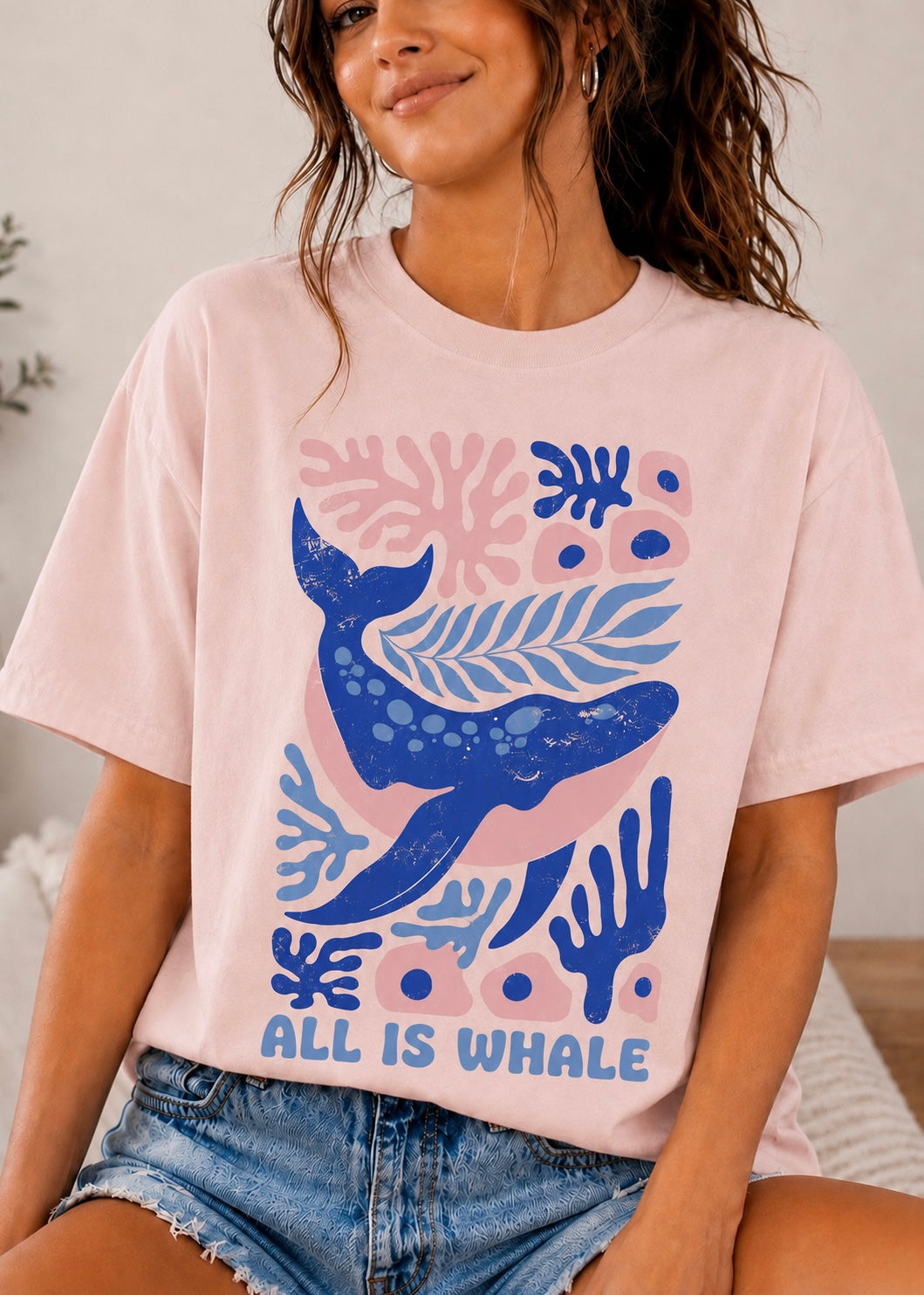 A woman wearing a light pink graphic t-shirt with a blue whale and coral design, paired with denim shorts.