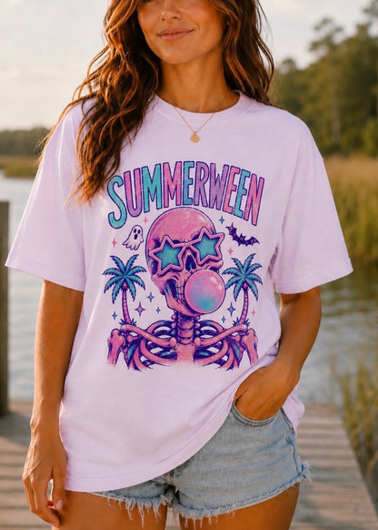 A woman wearing a light lavender graphic t-shirt with a purple and blue skull design and the word 'SUMMERWEEN' printed on the front, paired with denim shorts, standing outdoors near a body of water.