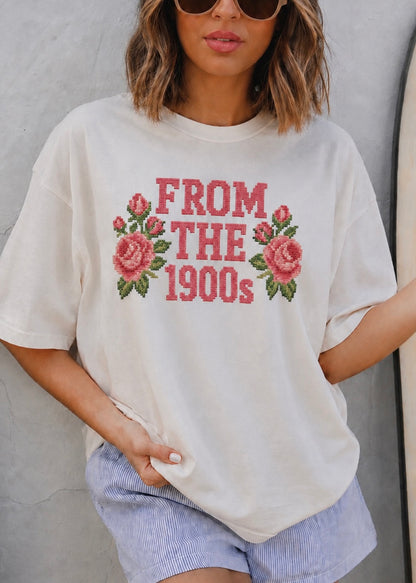 A woman wearing an oversized cream-colored graphic t-shirt with 'FROM THE 1900s' and pink roses printed on the front, paired with light blue striped shorts.