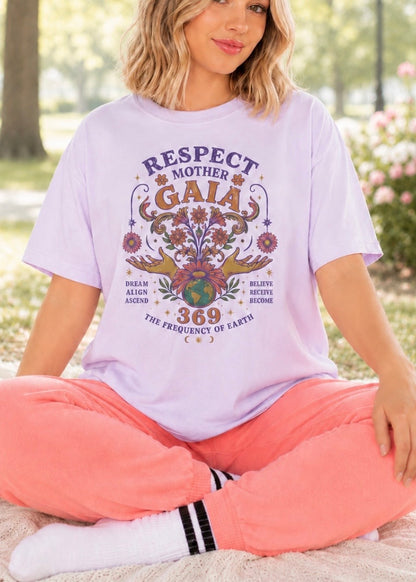 A woman sitting cross-legged outdoors wearing a light lavender graphic T-shirt with a colorful 'Respect Mother Gaia' design, paired with coral pink sweatpants and white socks.