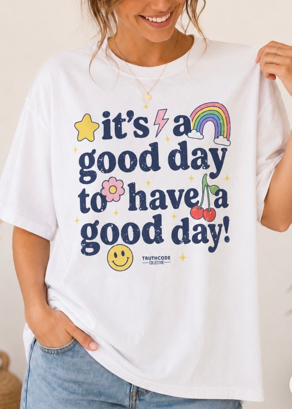 Person wearing a cute white t-shirt with colorful text 'It's a good day to have a good day' and star, rainbow, cherries, smiley face and flower graphics.