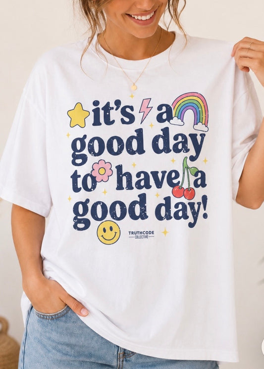 Person wearing a cute white t-shirt with colorful text 'It's a good day to have a good day' and star, rainbow, cherries, smiley face and flower graphics.