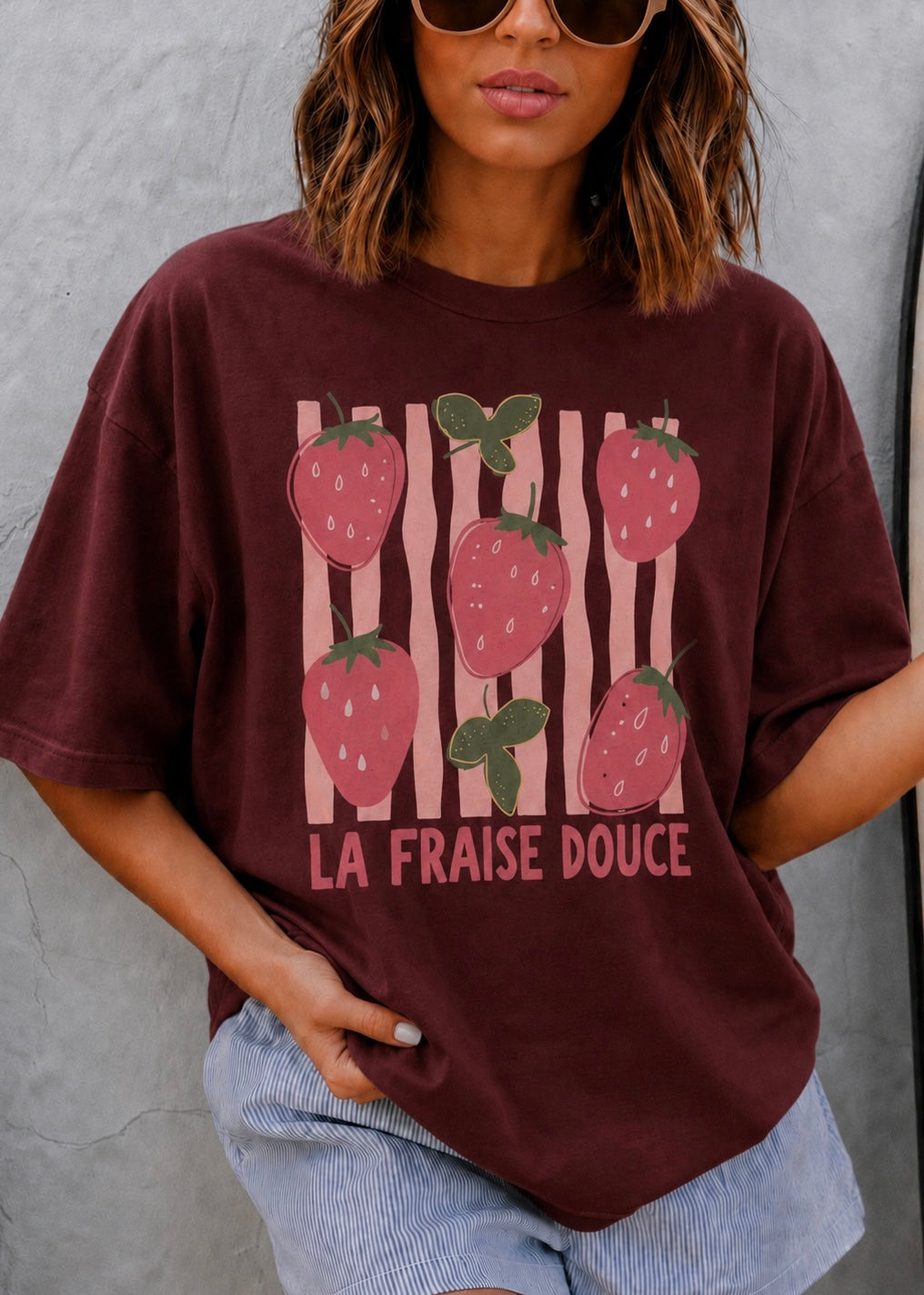 A woman wearing an oversized maroon graphic t-shirt with a strawberry print and the French text 'LA FRAISE DOUCE' on the front, paired with light blue denim shorts against a gray concrete wall.