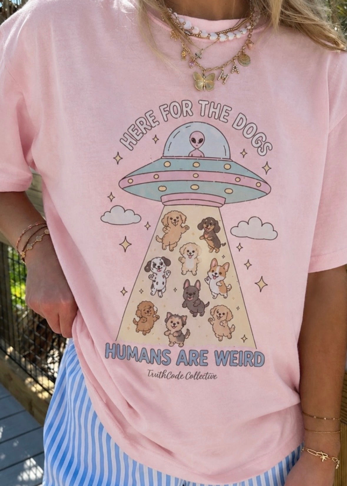 Person wearing a pink t-shirt with a cute UFO and dog illustration, text 'Here for the dogs humans are weird'.
