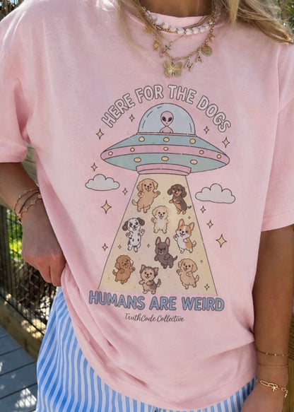Person wearing a pink t-shirt with a cute UFO and dog illustration, text 'Here for the dogs humans are weird'.