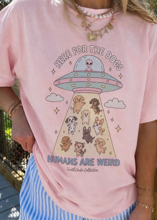 Person wearing a pink t-shirt with a cute UFO and dog illustration, text 'Here for the dogs humans are weird'.